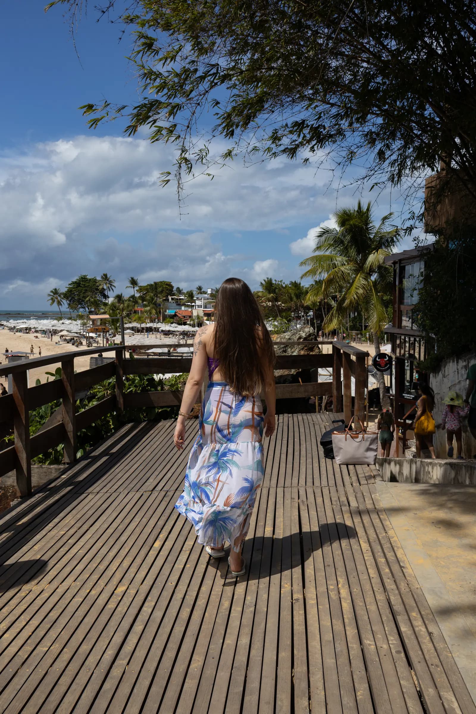 Mulher de vestido estampado caminha por uma passarela de madeira com vista para a praia