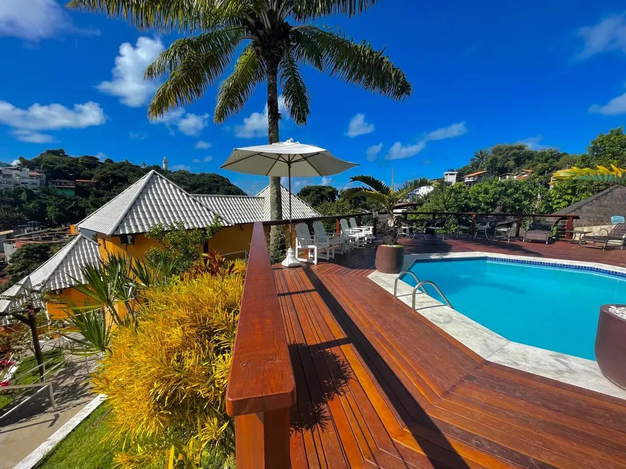 Piscina na cobertura do hotel, com deck de madeira, guarda-sol branco, espreguiçadeiras e vista para os telhados da vila