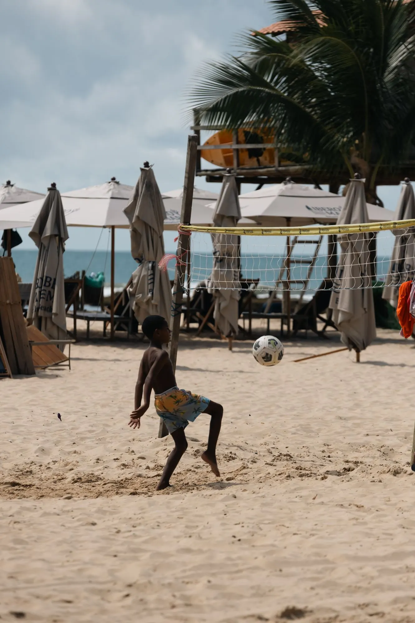 Menino joga bola na areia perto da rede de vôlei, com guarda-sóis, coqueiro e o mar ao fundo