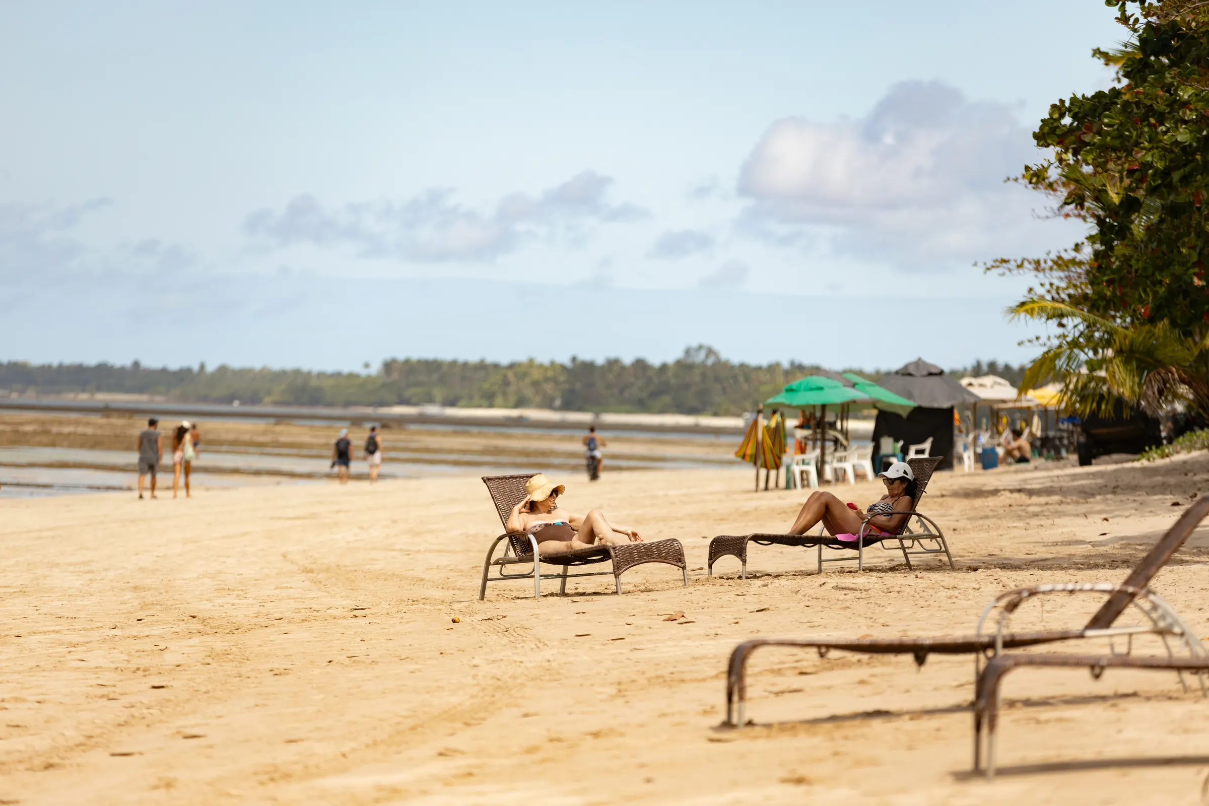 Praia de areia clara com espreguiçadeiras e pessoas caminhando na maré baixa