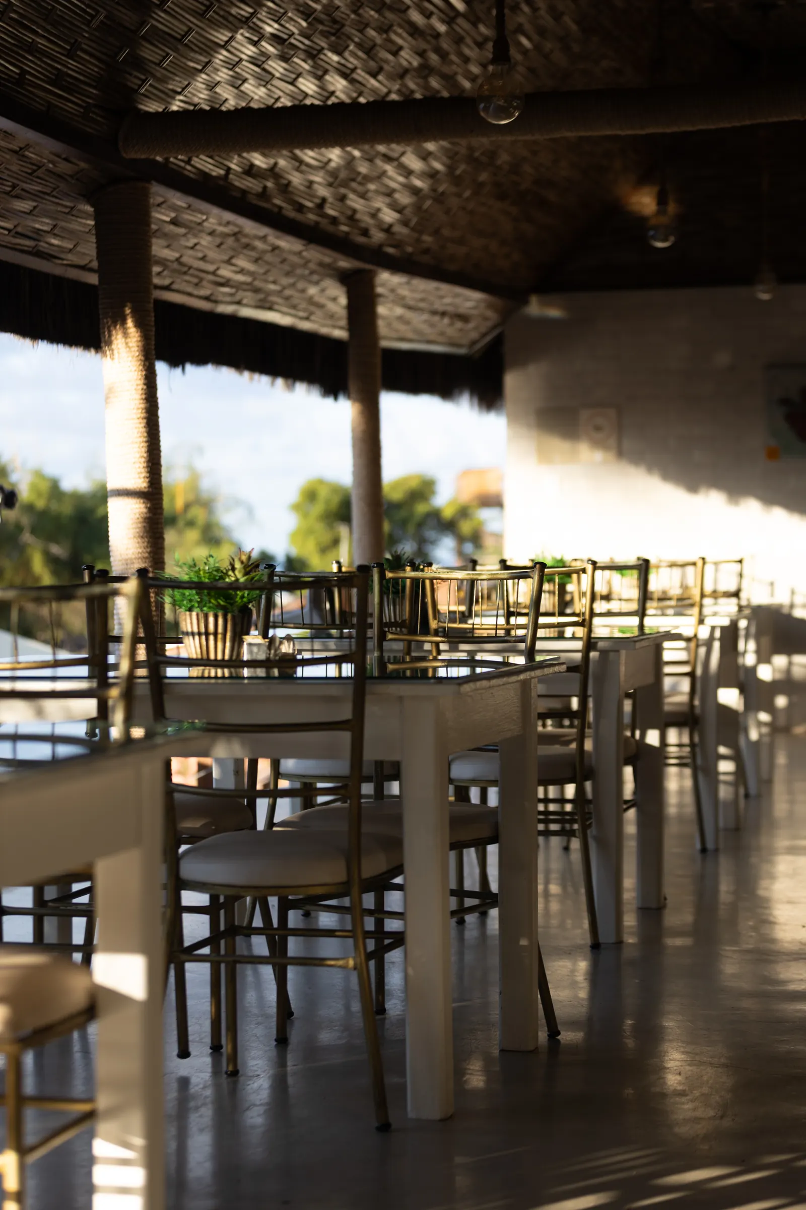 Salão do restaurante com cobertura de palha, mesas e cadeiras e luz da tarde